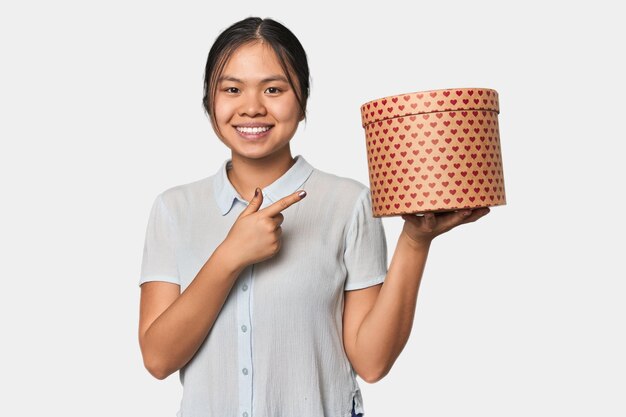 Молодая китайская женщина с коробкой, наполненной сердцем, улыбается и указывает в сторону, показывая что-то.