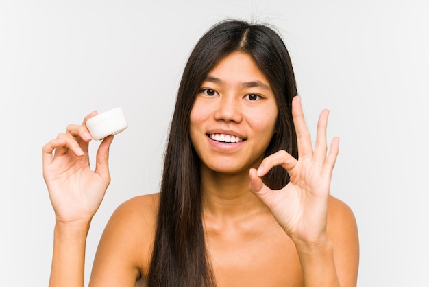 Young chinese woman holding a moisturizer isolated cheerful and confident showing ok gesture.