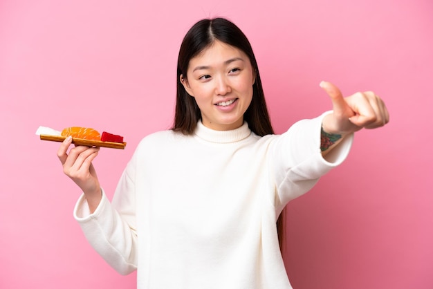 Young chinese woman holding sashimi isolated on pink background giving a thumbs up gesture