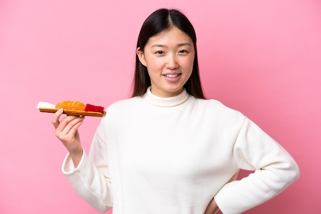 Young Chinese woman holding sashimi isolated on pink background posing with arms at hip and smiling