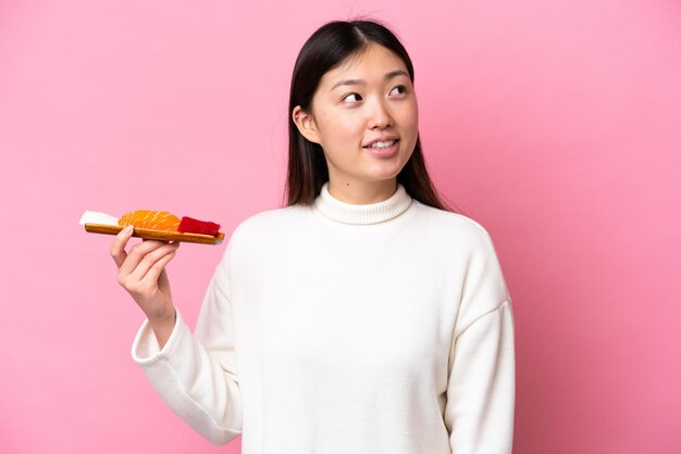 Young Chinese woman holding sashimi isolated on pink background thinking an idea while looking up