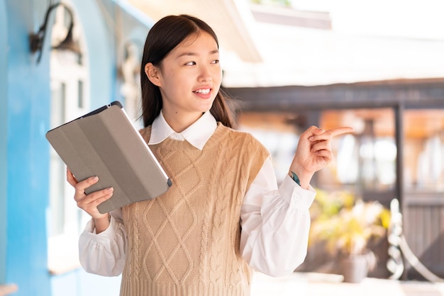 Young Chinese woman holding a tablet at outdoors pointing to the side to present a product