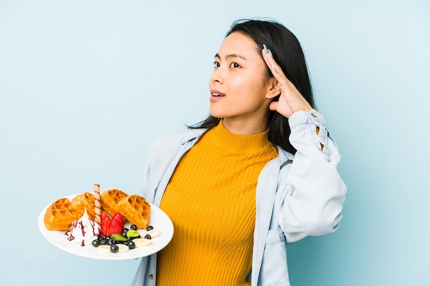 Young chinese woman holding waffle isolated on blue space  being shocked, she has remembered important meeting.