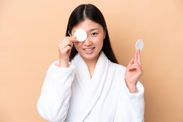 Young Chinese woman isolated on beige background with cotton pad for removing makeup from her face and smiling