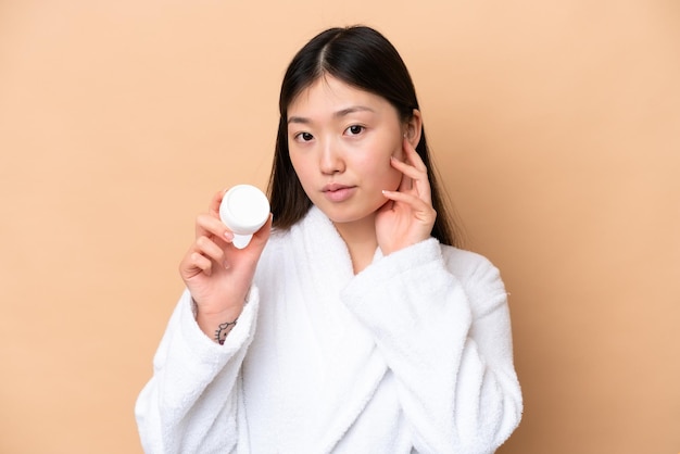 Young Chinese woman isolated on beige background with moisturizer