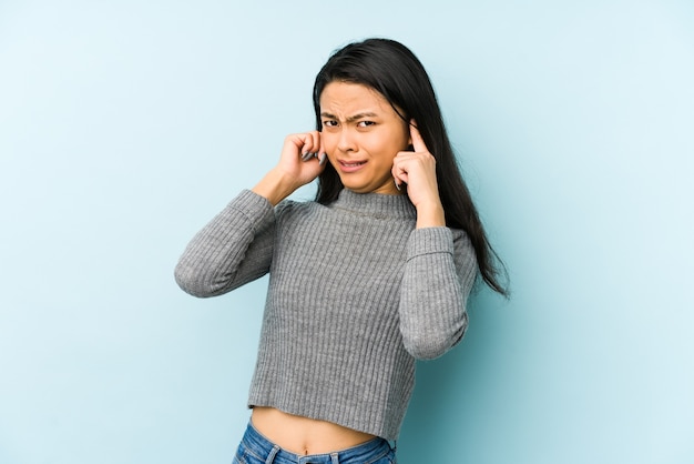 Young chinese woman isolated on a blue wall covering ears with hands.