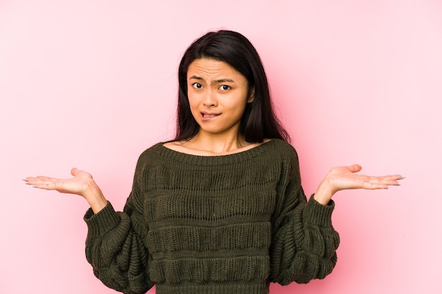 Photo young chinese woman isolated on a pink background confused and doubtful shrugging shoulders to hold a copy space.