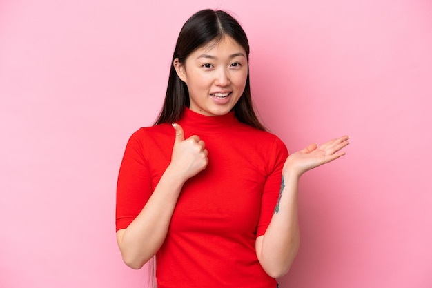 Young Chinese woman isolated on pink background holding copyspace imaginary on the palm to insert an ad and with thumbs up