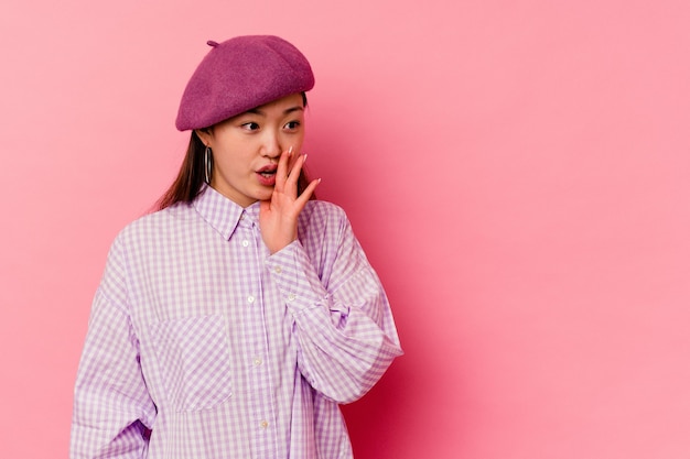 Young chinese woman isolated on pink background is saying a secret hot braking news and looking aside