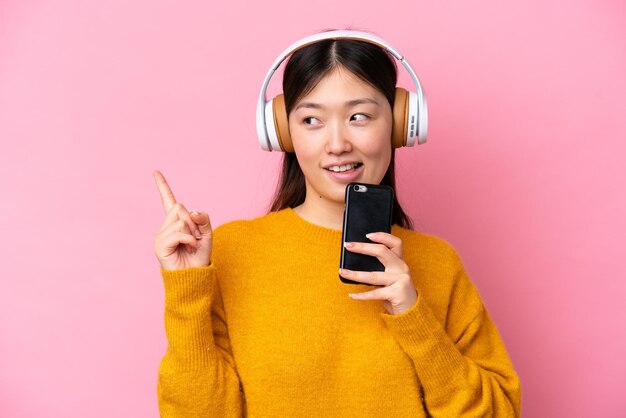 携帯電話で音楽を聴いて歌うピンクの背景に分離された若い中国人女性