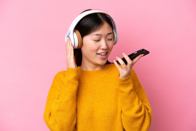 Giovane donna cinese isolata su sfondo rosa che ascolta musica con un cellulare e canta