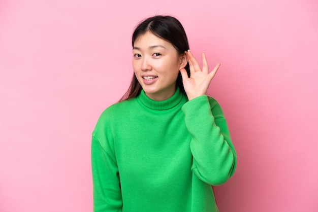 Young Chinese woman isolated on pink background listening to something by putting hand on the ear