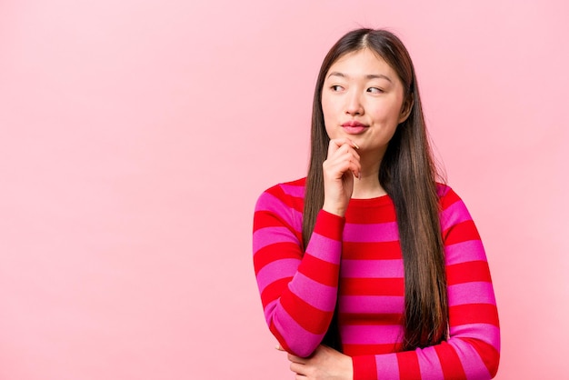 Young Chinese woman isolated on pink background relaxed thinking about something looking at a copy space