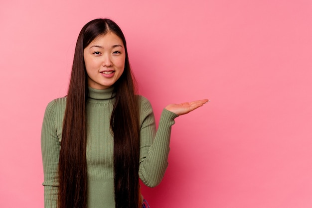 Young chinese woman isolated on pink background showing a copy space on a palm and holding another hand on waist.