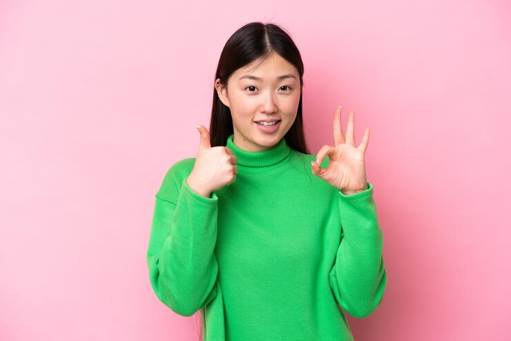 Photo ピンクの背景に分離された若い中国人女性 ok のサインと親指アップ ジェスチャーを示す