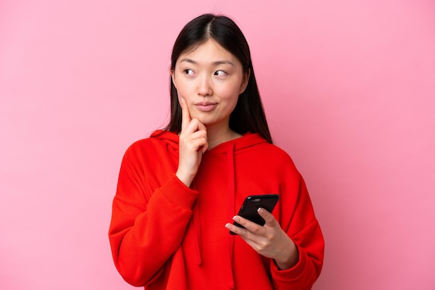 Young chinese woman isolated on pink background using mobile phone and thinking