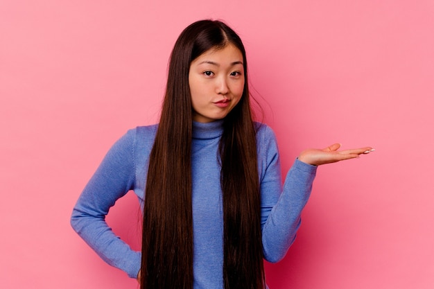 Young chinese woman isolated on pink wall doubting and shrugging shoulders in questioning gesture.