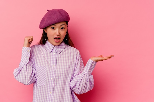 Young chinese woman isolated on pink wall holds copy space on a palm, keep hand over cheek. Amazed and delighted.