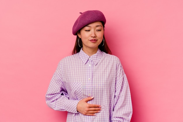 Young chinese woman isolated on pink wall touches tummy, smiles gently, eating and satisfaction concept.