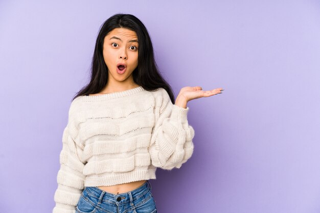 Young chinese  woman isolated on a purple wall impressed holding copy space on palm.