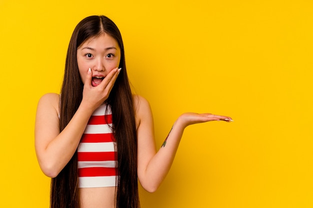 Young chinese woman isolated on yellow background holds copy space on a palm, keep hand over cheek. Amazed and delighted.