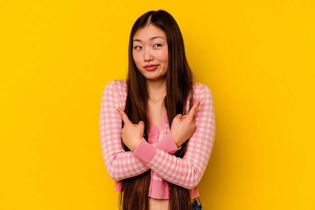 Young chinese woman isolated on yellow background points sideways, is trying to choose between two options.