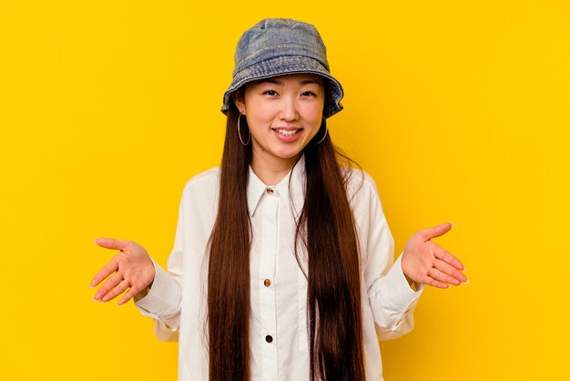 Young chinese woman isolated on yellow holding something with both hands, product presentation.