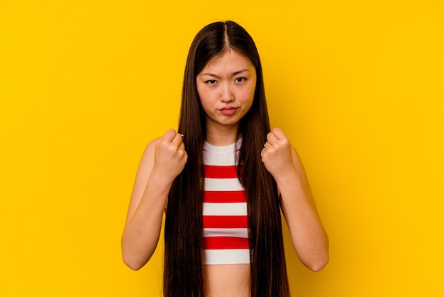 Young chinese woman isolated on yellow showing fist, aggressive facial expression.