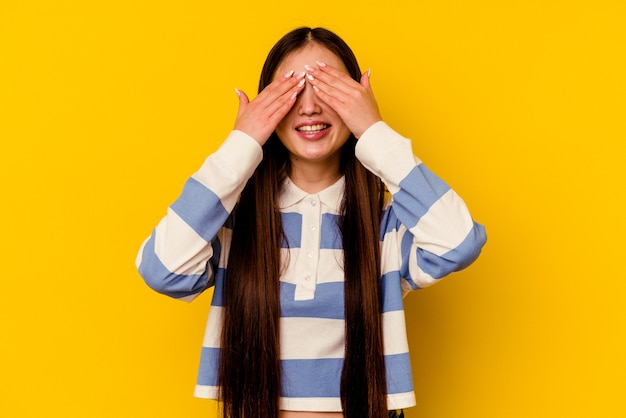 young chinese woman isolated on yellow wall covers eyes with hands, smiles broadly waiting for a surprise