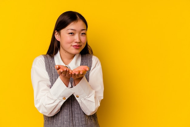 Young chinese woman isolated on yellow wall folding lips and holding palms to send air kiss