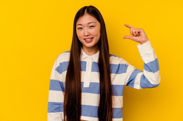 Молодая китаянка изолирована на желтой стене, держа что-то немного указательными пальцами, улыбаясь и уверенно.
