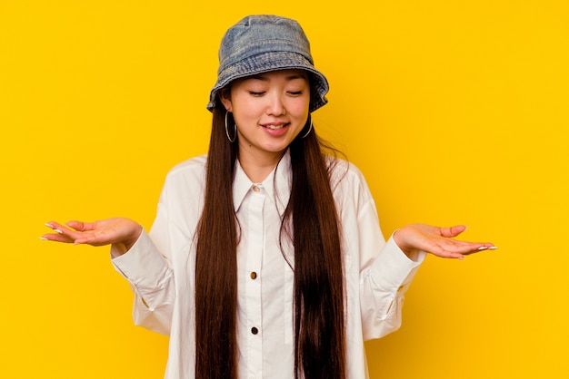 Young chinese woman isolated on yellow wall holding something with palms, offering to front