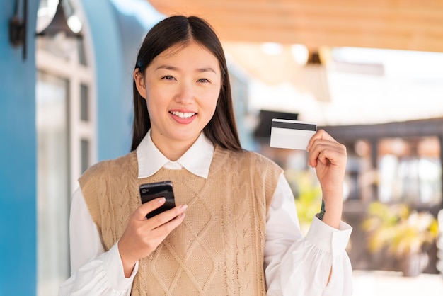 Giovane donna cinese all'aperto che compra con il cellulare con una carta di credito