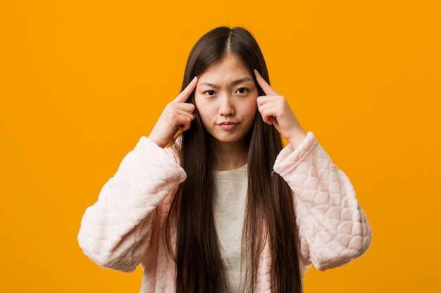 Young chinese woman in pajama focused on a task, keeping him forefingers pointing head.