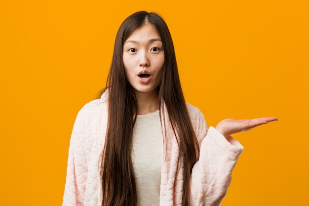 Young chinese woman in pajama impressed holding copy space on palm.