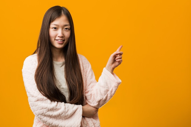 Young chinese woman in pajama smiling cheerfully pointing with forefinger away.