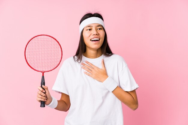 Young chinese woman playing badminton in a pink wall laughs out loudly keeping hand on chest.