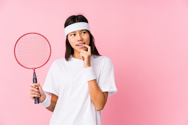 Young chinese woman playing badminton in a pink wall relaxed thinking about something looking at a blank space.