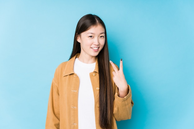 Молодая китаянка позирует в синем фоне изолированы, показывая знак победы и широко улыбаясь.