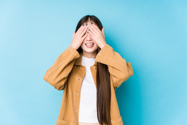 Young chinese woman posing in a blue space isolated covers eyes with hands, smiles broadly waiting for a surprise.