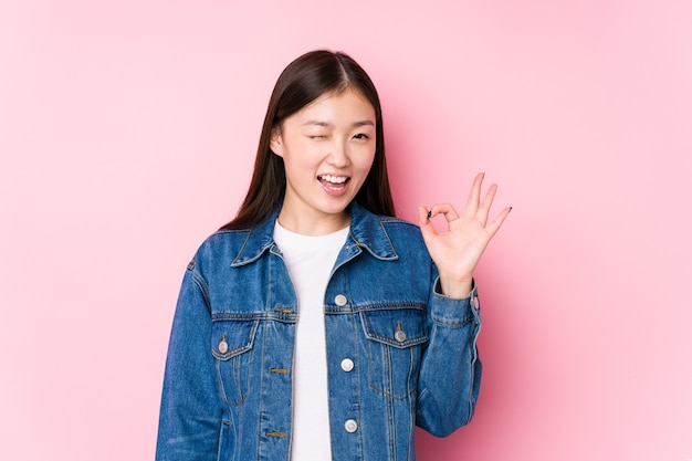 Young chinese woman posing in a pink background isolated winks an eye and holds an okay gesture with hand.