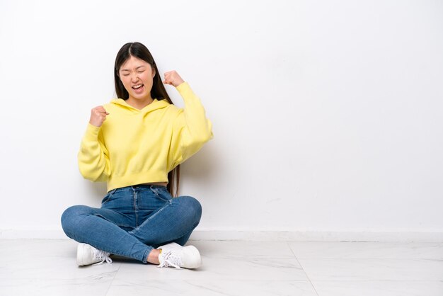 Young Chinese woman sitting on the floor isolated on white wall celebrating a victory