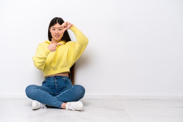 Молодая китаянка сидит на полу, изолированная на белой стене, фокусируясь на символе кадрирования лица