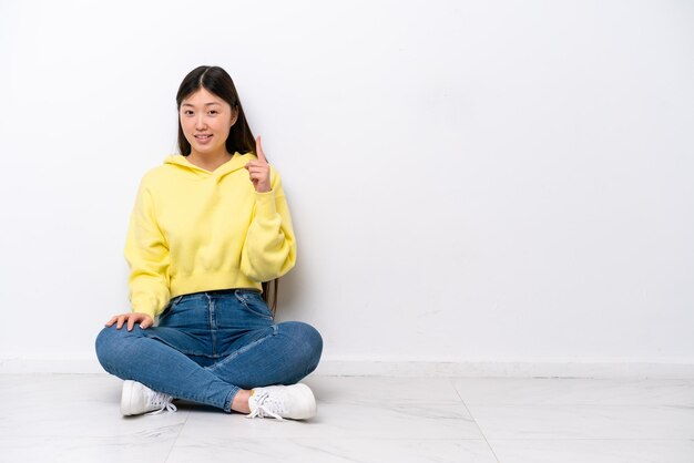 Young Chinese woman sitting on the floor isolated on white wall pointing with the index finger a great idea