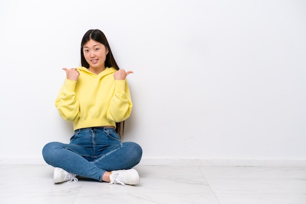 Young Chinese woman sitting on the floor isolated on white wall with thumbs up gesture and smiling