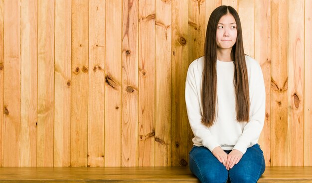 Young chinese woman sitting on a wooden place confused, feels doubtful and unsure.