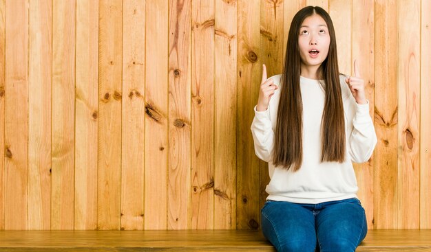 Young chinese woman sitting on a wooden place pointing upside with opened mouth.