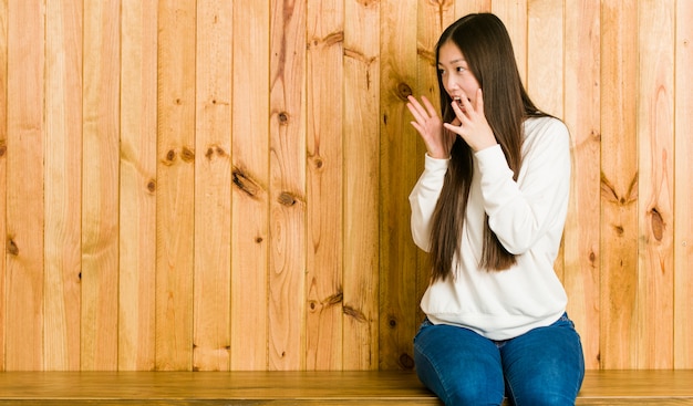 Young chinese woman sitting on a wooden place shouts loud, keeps eyes opened and hands tense.