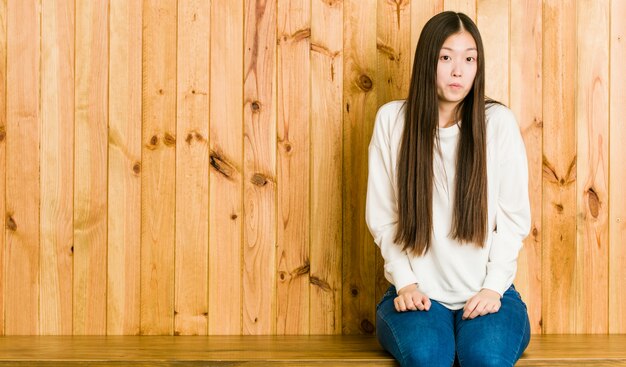 Young chinese woman sitting on a wooden place shrugs shoulders and open eyes confused.