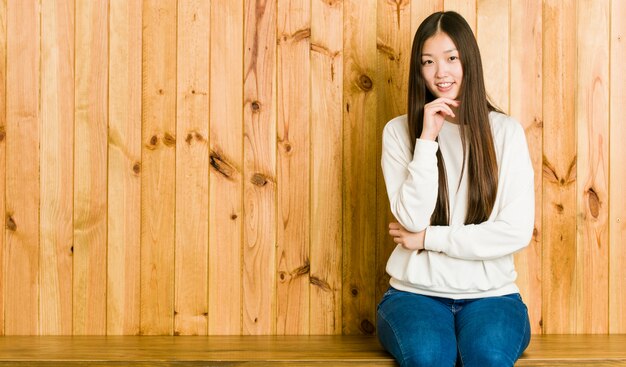 Young chinese woman sitting on a wooden place smiling happy and confident, touching chin with hand.